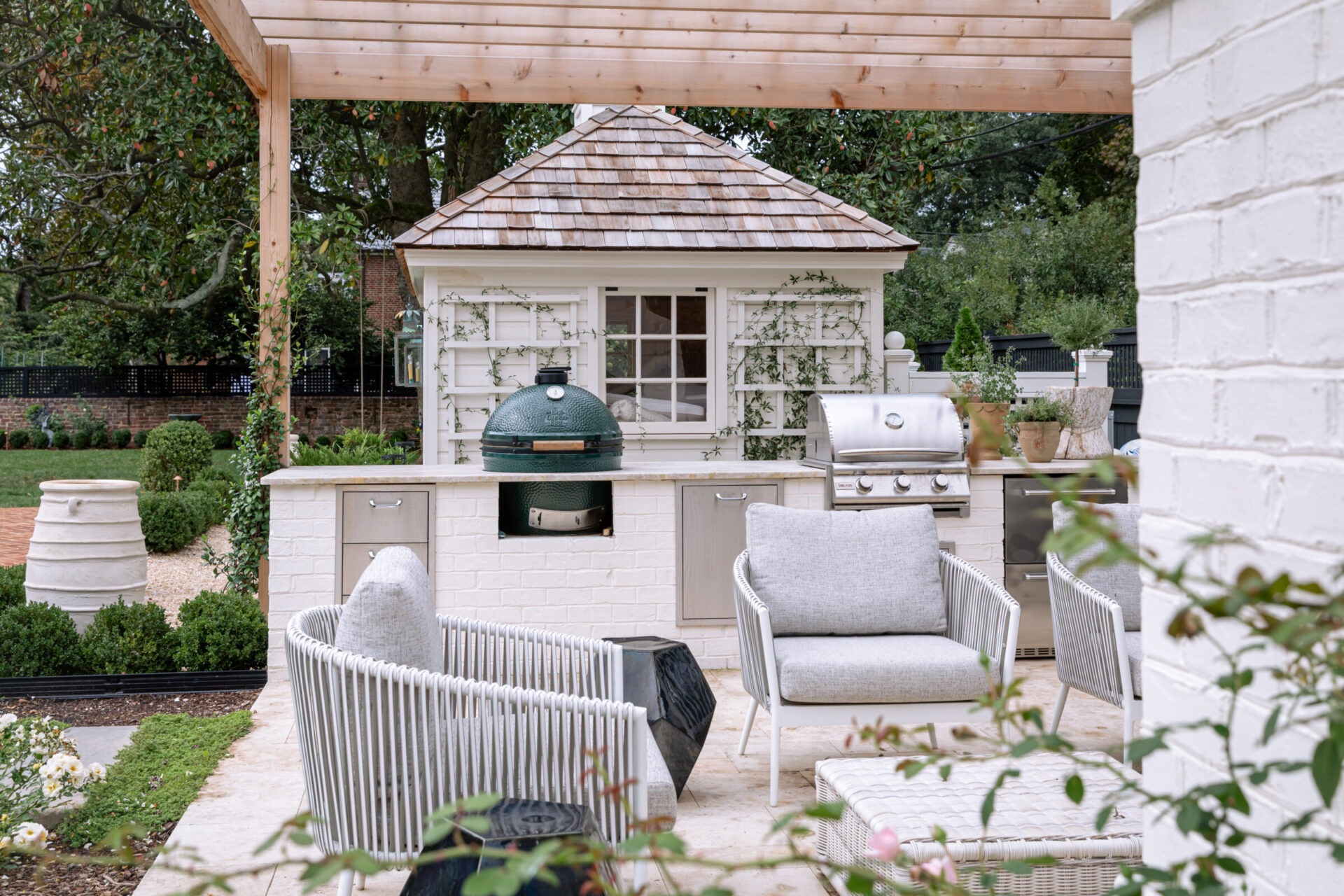 Outdoor patio kitchen features comfortable seating, a grill, and a green egg smoker under a wooden pergola, surrounded by lush greenery and garden.