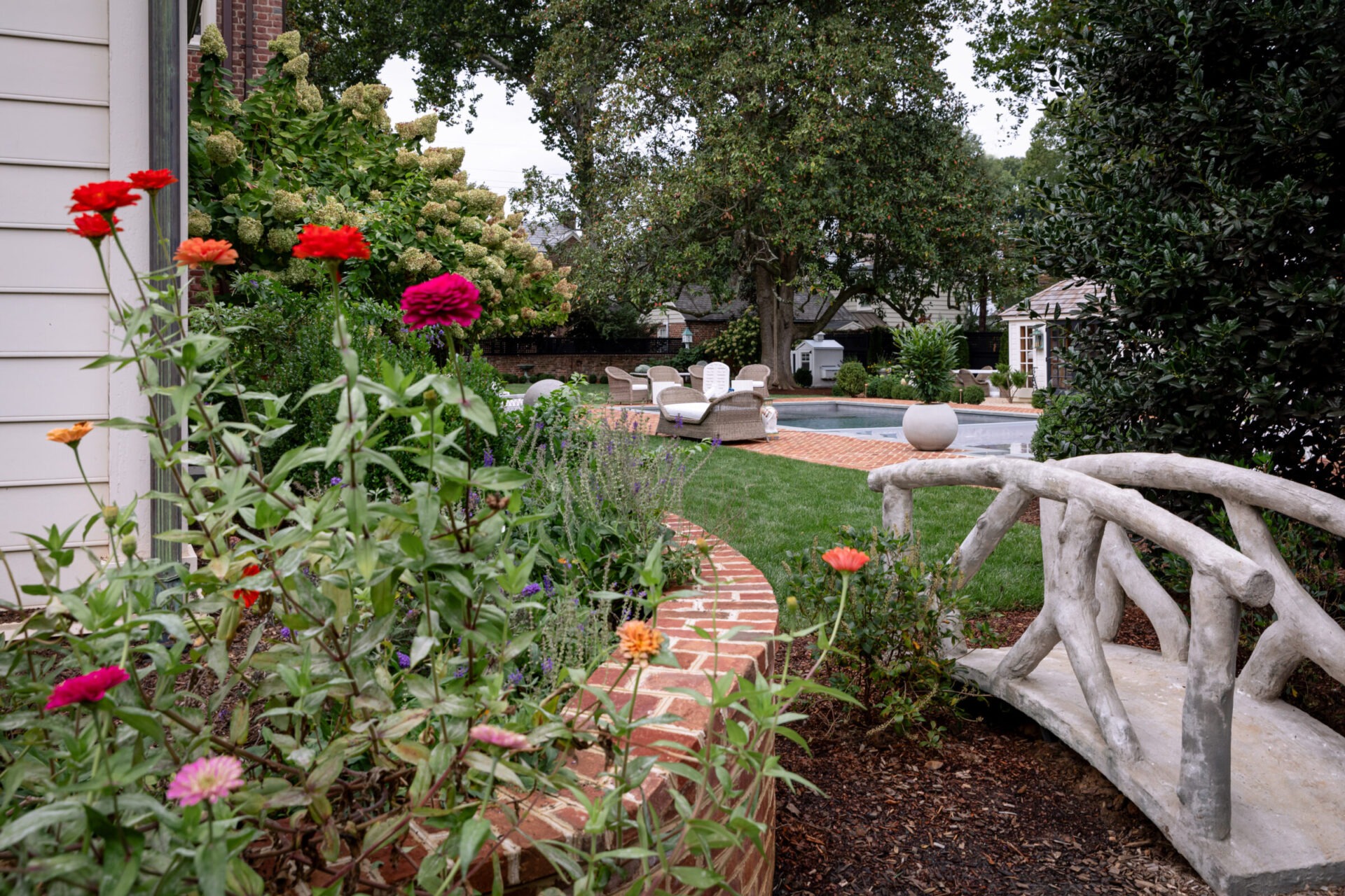 A garden scene features colorful flowers, a small curved bridge, and a pool area with lounge chairs surrounded by lush greenery and trees.