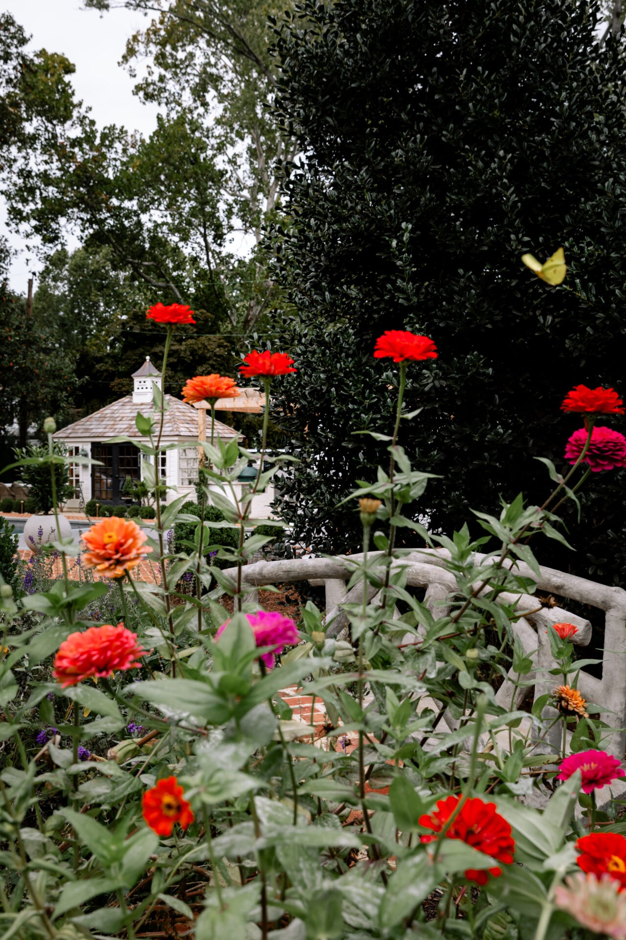 Colorful flowers in a lush garden with a butterfly, stone railing, and a gazebo in the background. Trees surround this tranquil setting.