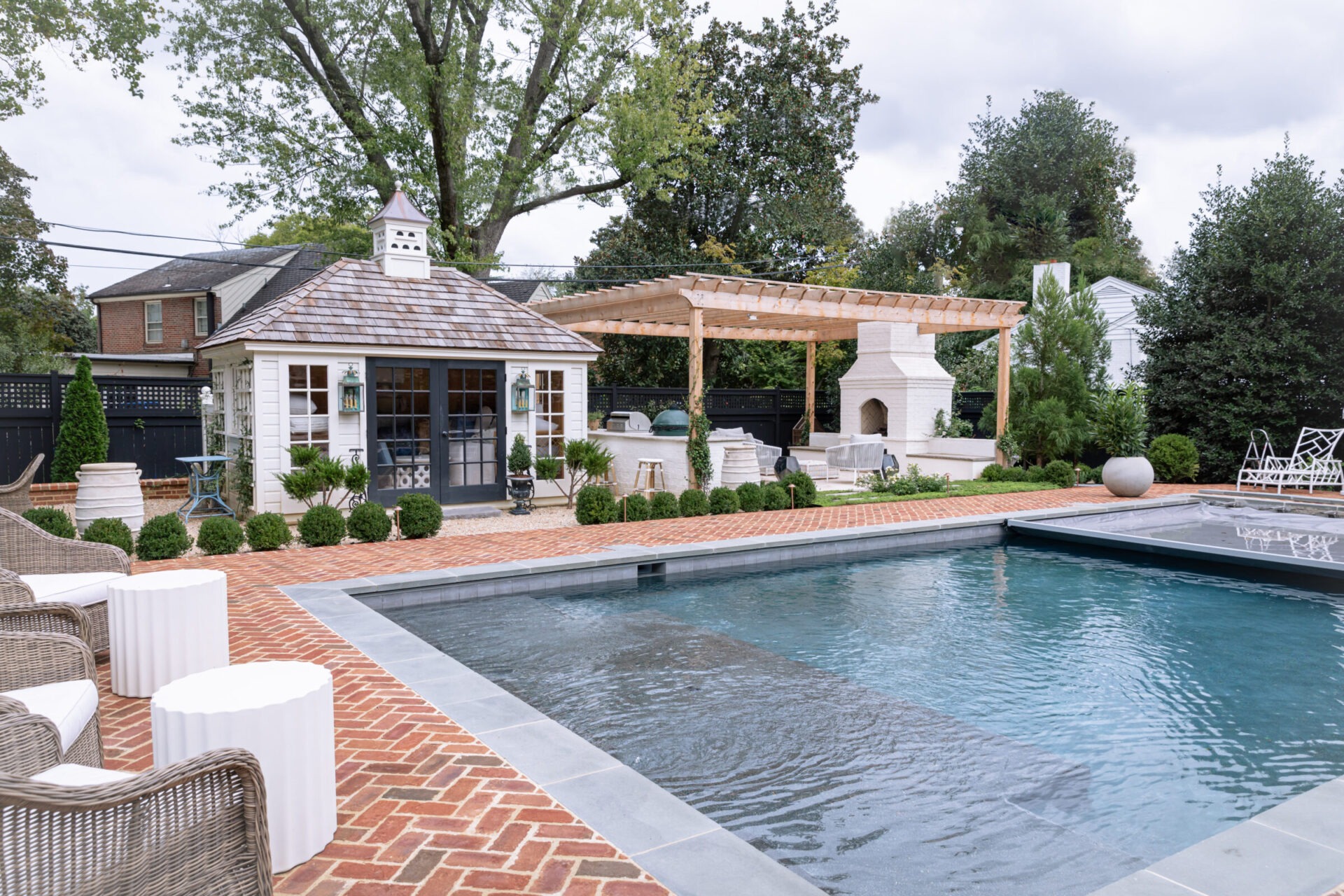 Charming backyard features a pool, brick patio, pergola, and small house. Lush trees and greenery surround the serene, cozy setting.