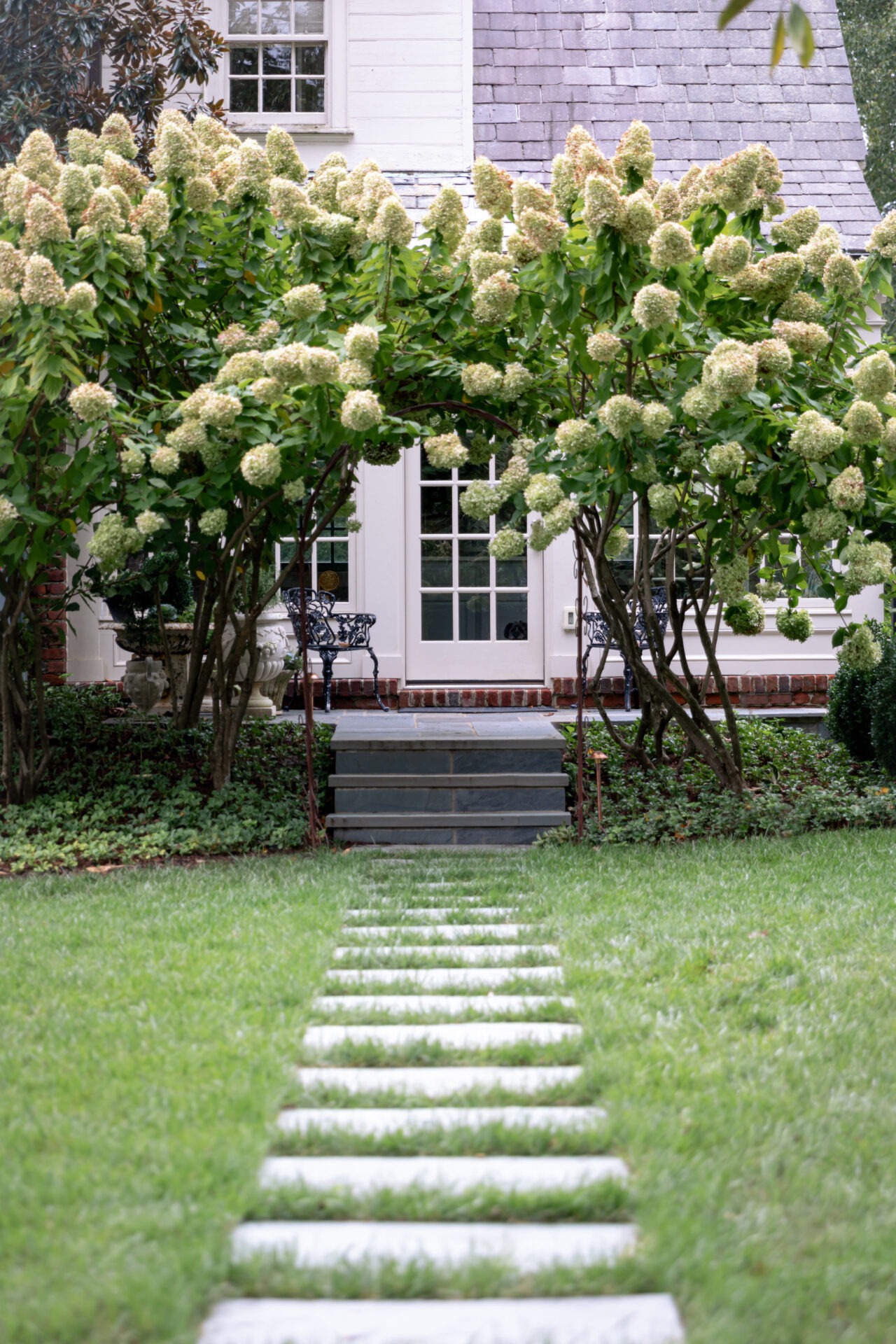 A charming house entrance framed by lush hydrangea bushes, stone path leads to door, surrounded by green lawn and outdoor seating.