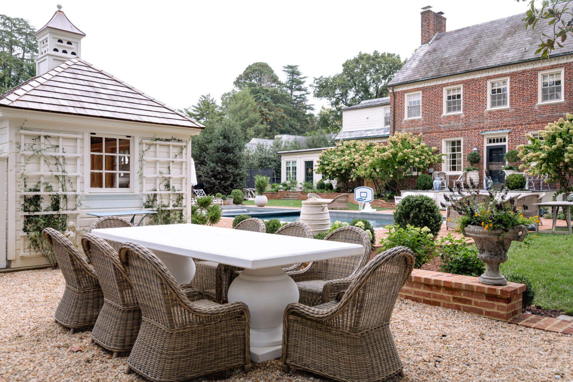 A quaint, historic brick house with a charming backyard patio, wicker chairs, white gazebo, lush greenery, and a cozy outdoor dining area.