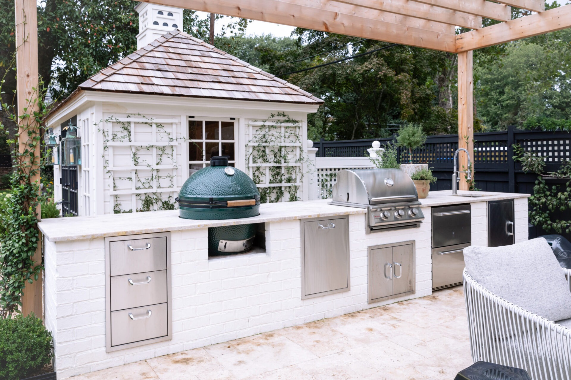 Outdoor kitchen with grill and green ceramic cooker under a wooden pergola. Small white garden shed with trellis in background. Lush greenery surrounds.