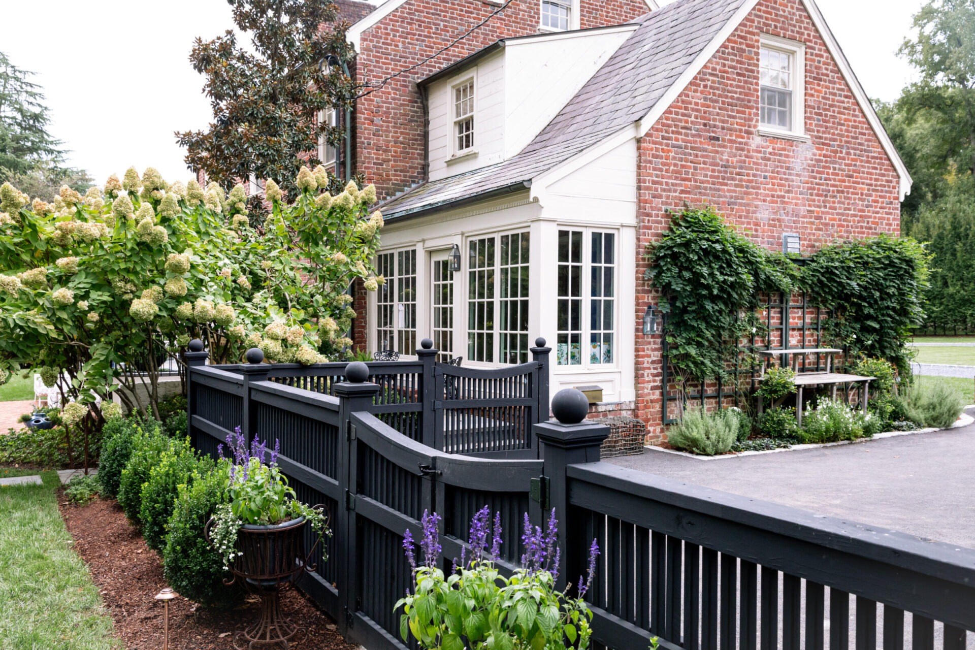 Charming brick house with a white-trimmed sunroom, surrounded by lush greenery and a wooden fence. Purple flowers line the garden path.