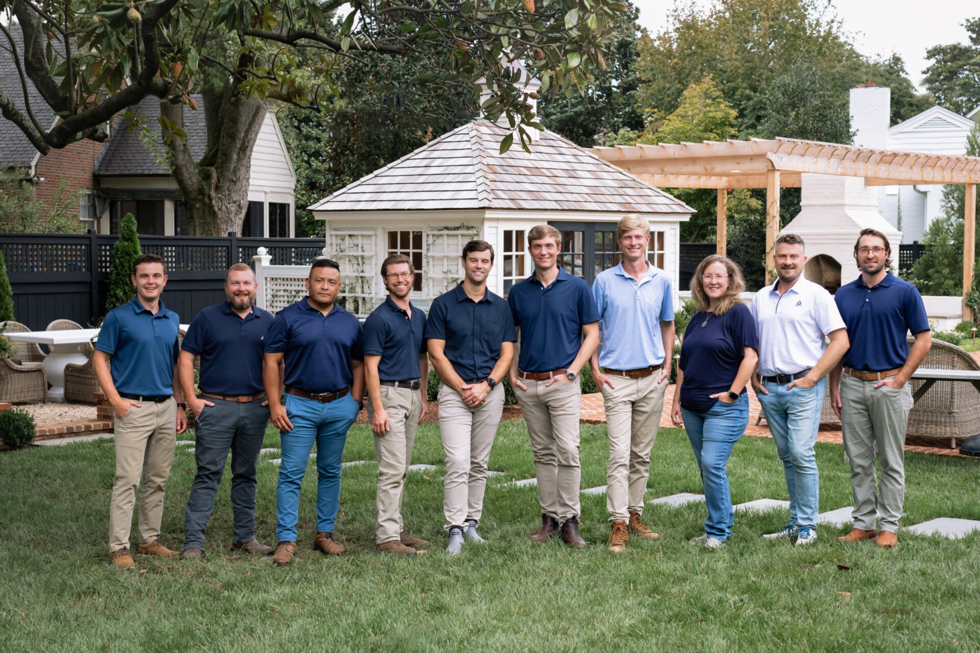 A group of people standing on grass in a garden setting with a small pavilion and trees in the background.