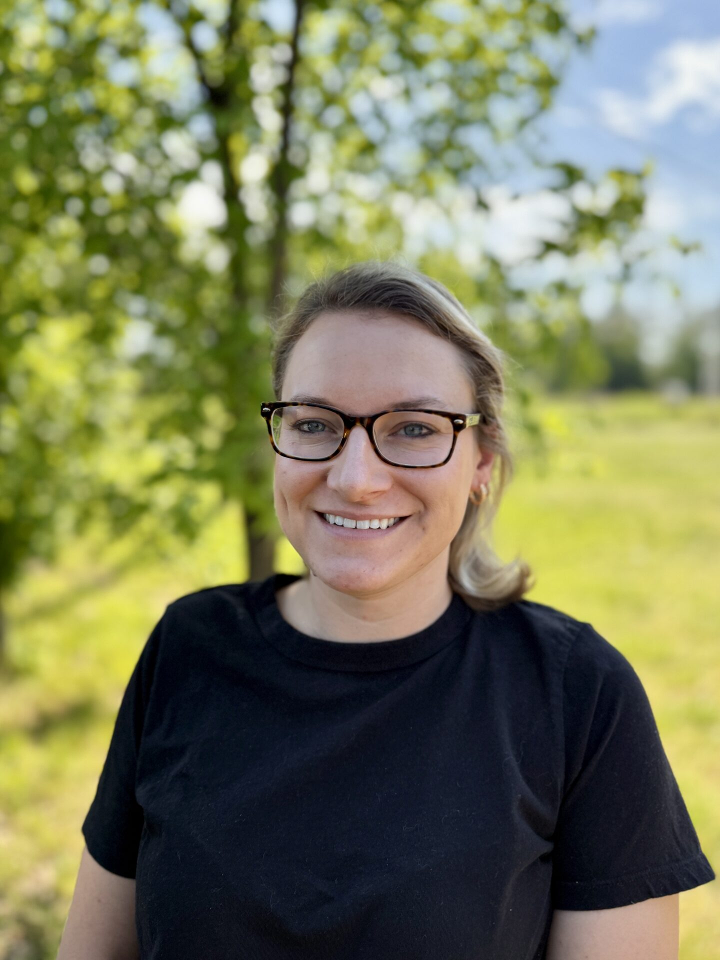 A person wearing glasses and a black shirt smiles in a sunny park with green trees and a blue sky in the background.