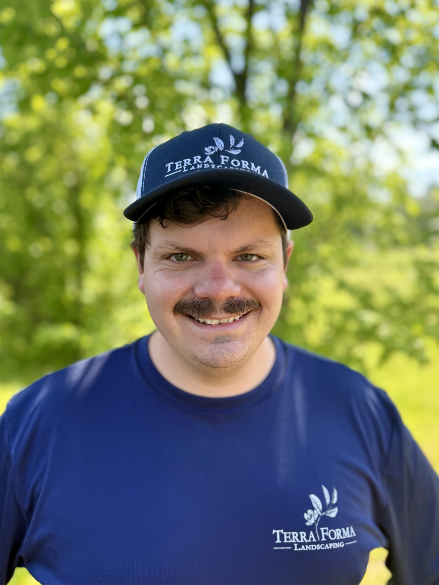 A person wearing a "Terra Forma Landscaping" cap and shirt smiles outdoors, surrounded by green trees and sunlight in the background.