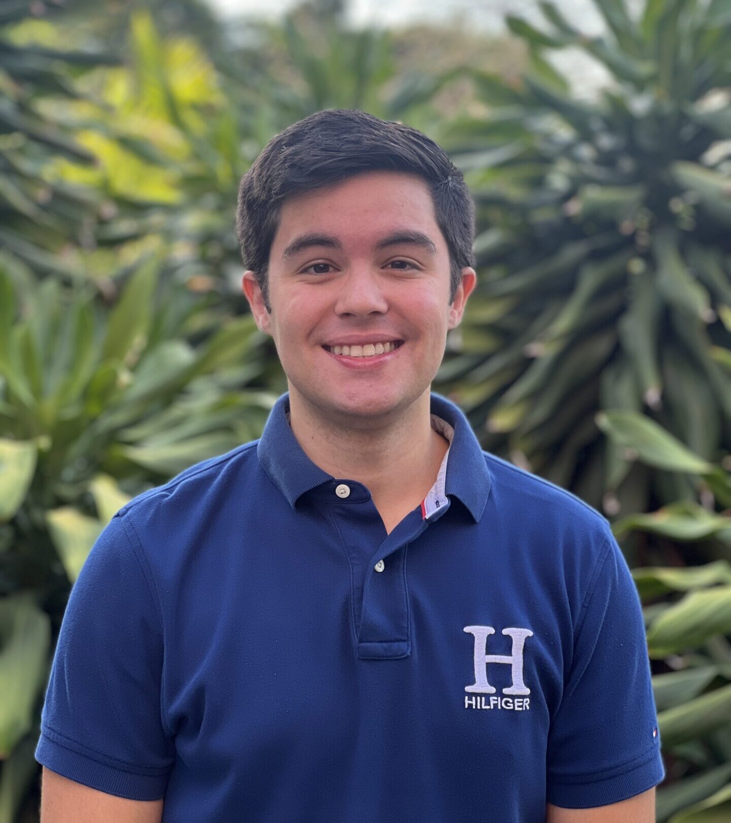 A person in a blue shirt stands smiling outdoors with lush green foliage in the background, under soft, natural lighting.