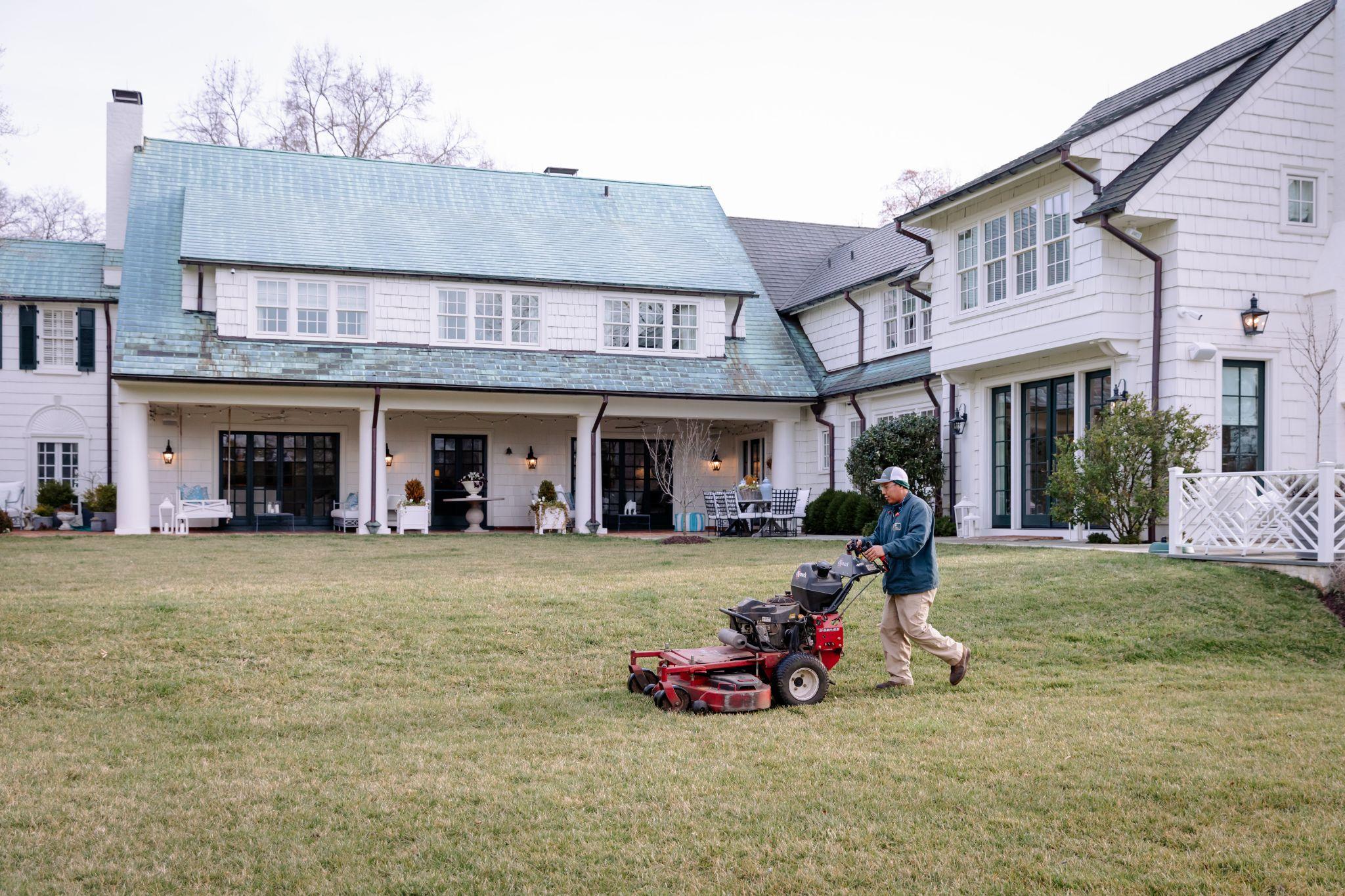 Lawn care professional mowing grass at a large white estate.